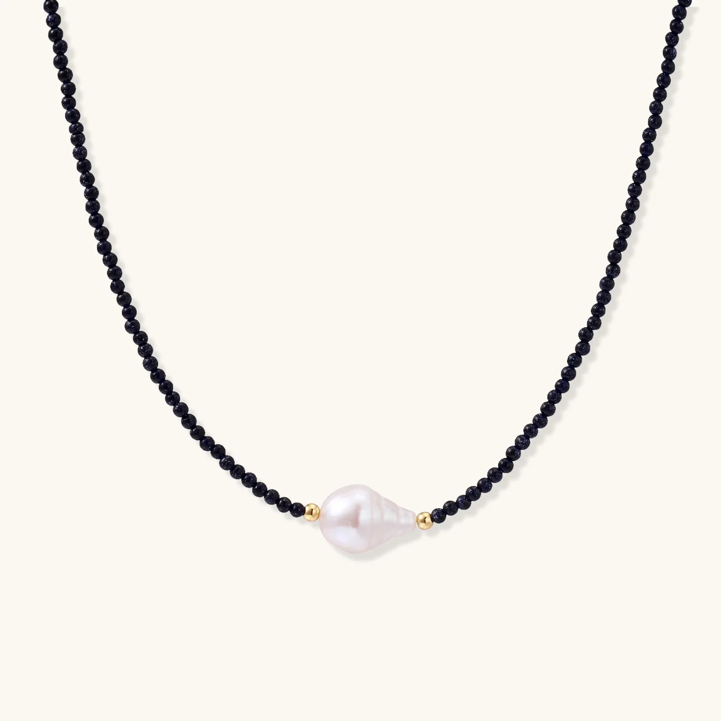 Delicate necklace with black blue sandstone beads and baroque pearl center on white background with gold accents.