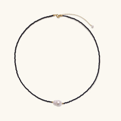 Delicate necklace with black blue sandstone beads and baroque pearl center on white background with gold accents.