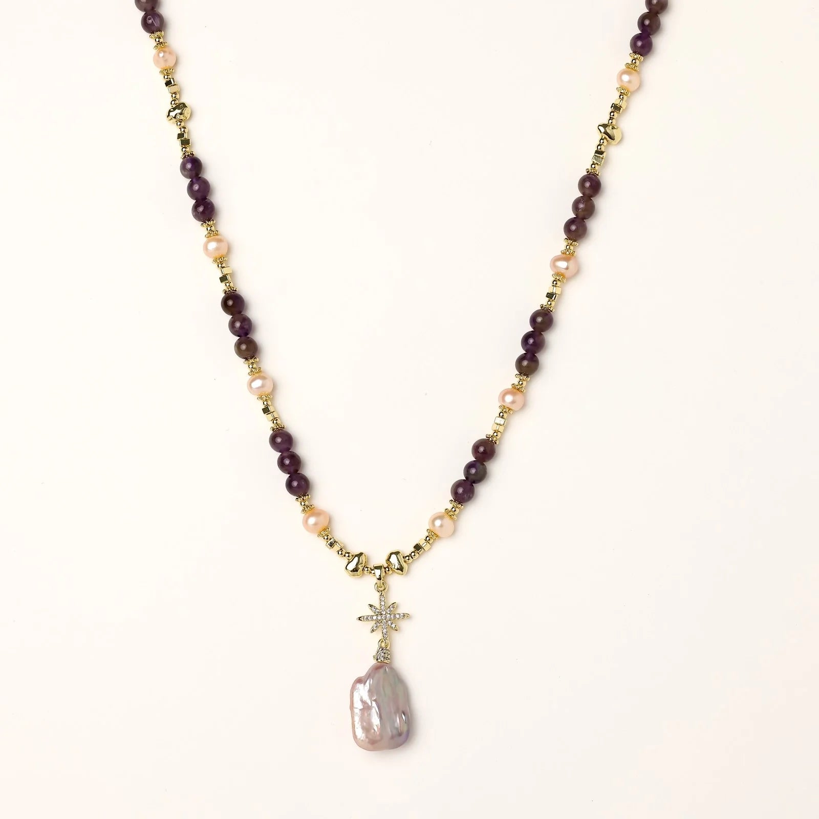 Gold necklace with iridescent baroque pearl pendant, purple amethyst beads, and crystal star charm on cream background