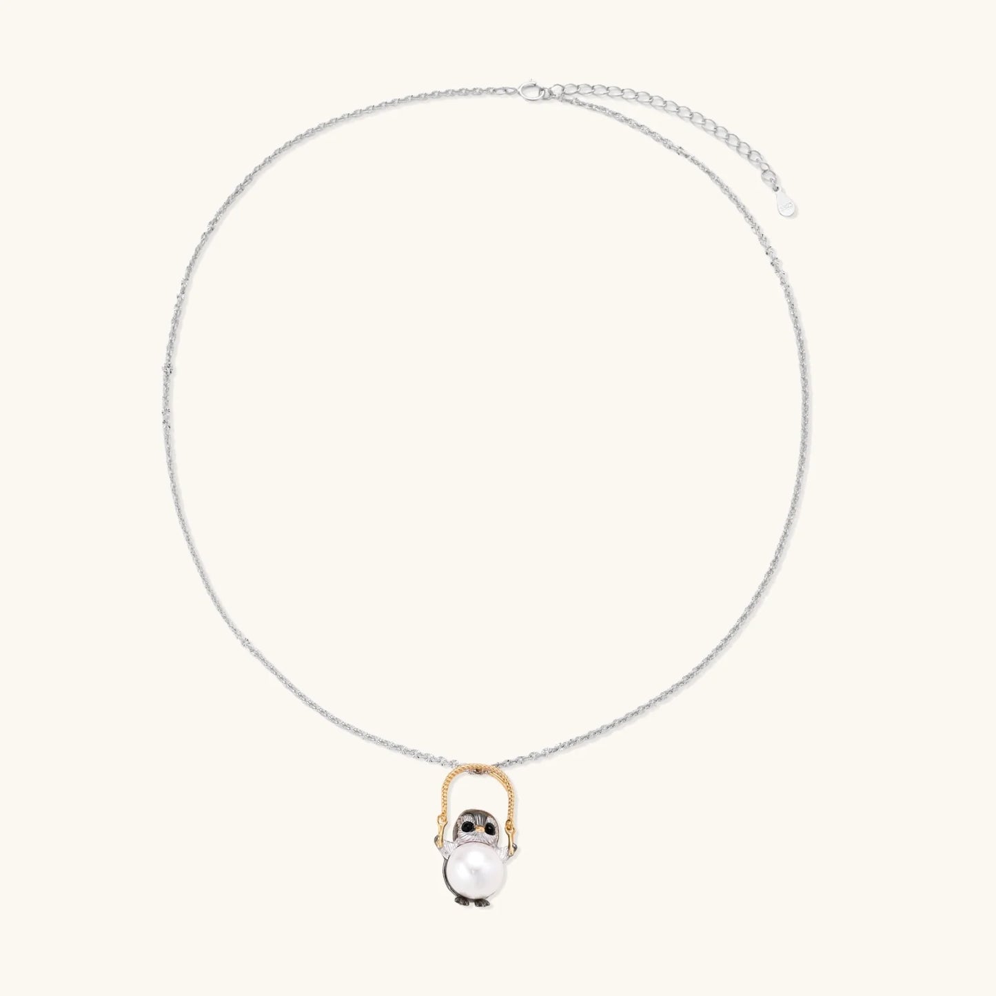 Whimsical owl pendant holding a white pearl on a gold swing, rhodium-plated silver chain on cream background.