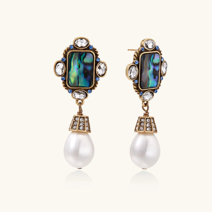 Ornate gold-plated earrings with iridescent abalone shell, crystals, blue stones, and white pearl drops on cream background.
